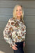 A smiling blonde woman wearing a cream-colored long-sleeve blouse with a detailed multicolor floral and botanical pattern, paired with dark jeans, standing against a gray wood-paneled wall.