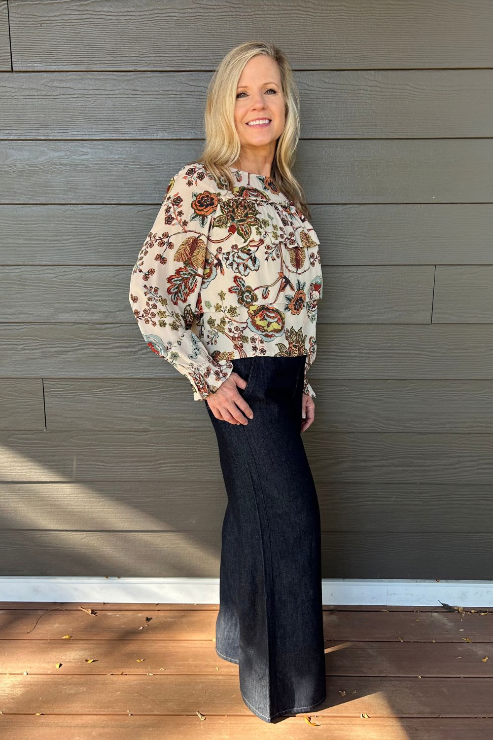 A woman stands on a wooden deck in front of a gray wood-paneled wall, wearing a long-sleeve cream blouse with a colorful floral and bird pattern, paired with a dark wide-leg skirt.