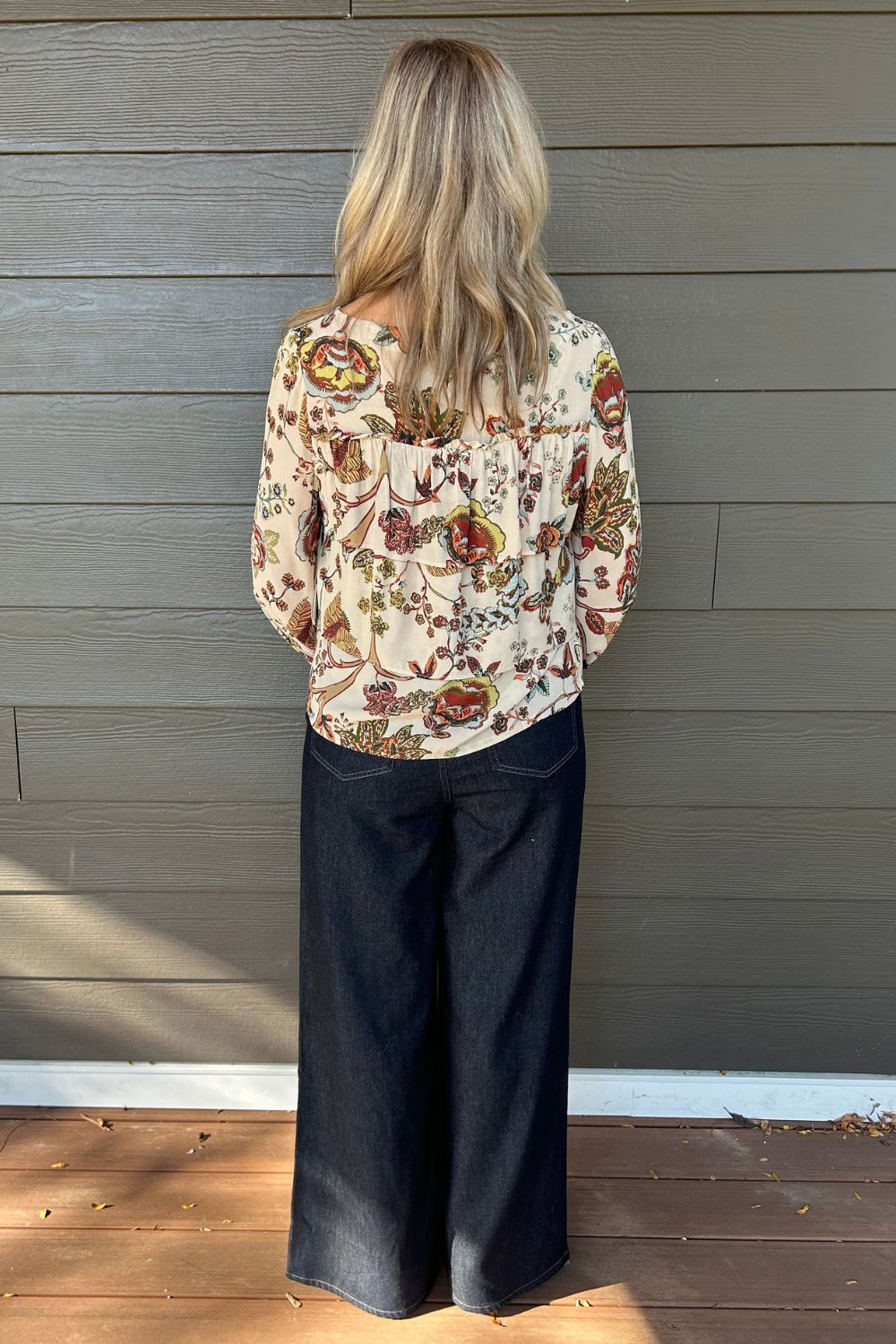 A woman standing with her back to the camera, wearing a cream-colored long-sleeve blouse with a colorful floral and botanical pattern, paired with dark wide-leg jeans, set against a gray wooden wall.