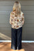 A woman standing with her back to the camera, wearing a cream-colored long-sleeve blouse with a colorful floral and botanical pattern, paired with dark wide-leg jeans, set against a gray wooden wall.