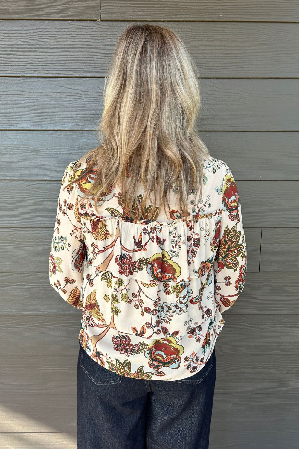 A woman viewed from behind wearing a cream-colored blouse with a dense, multicolor floral and botanical print in red, green, and orange, paired with dark blue jeans, standing against a gray wood-paneled wall.