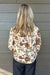 A woman viewed from behind wearing a cream-colored blouse with a dense, multicolor floral and botanical print in red, green, and orange, paired with dark blue jeans, standing against a gray wood-paneled wall.