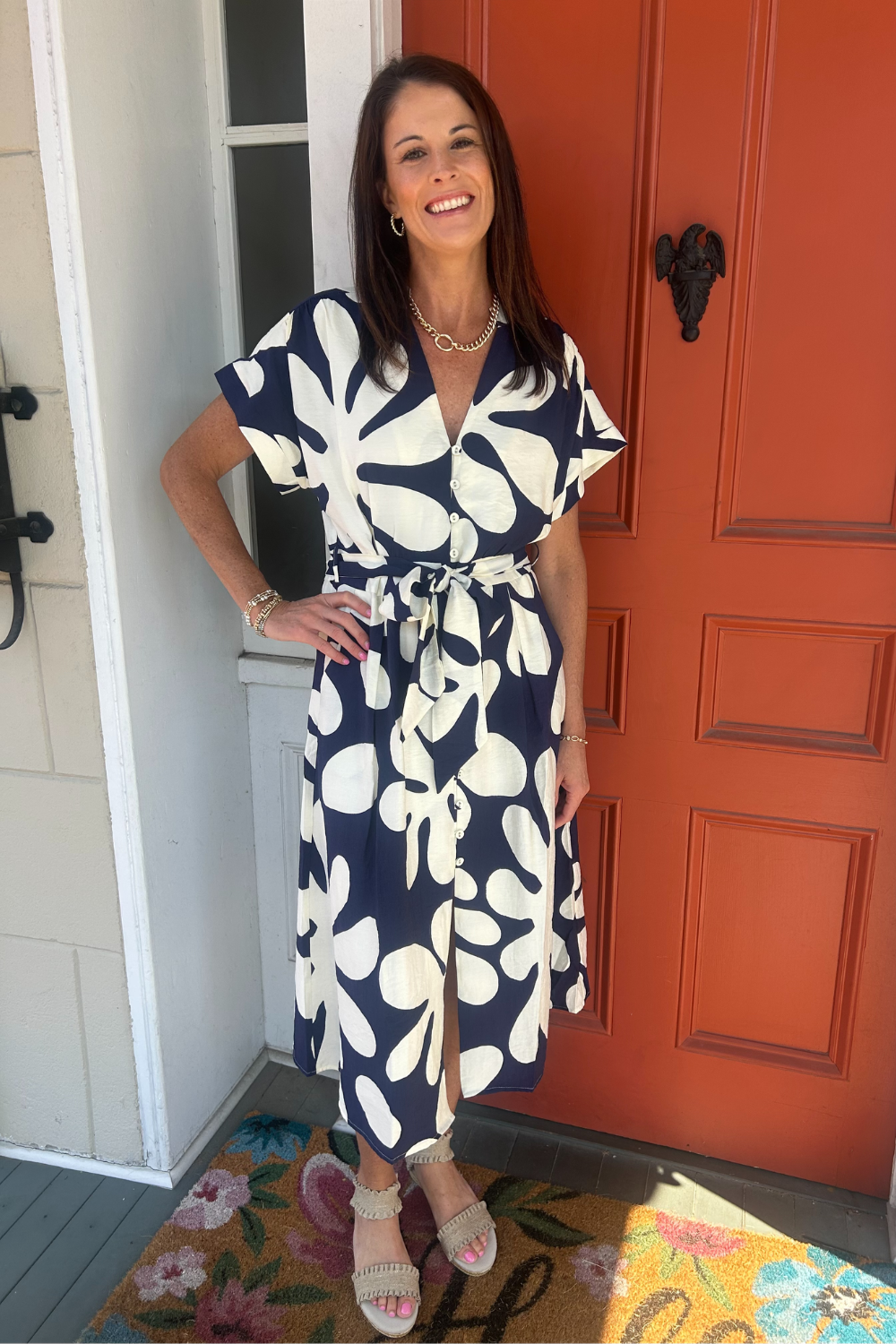 A woman wearing a navy and white floral print wrap dress with short flutter sleeves and a tie waist, standing on a porch in front of a bright orange door.