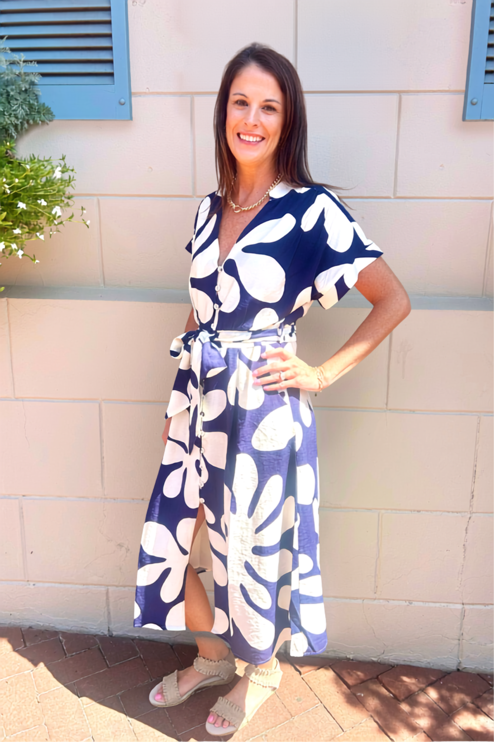 A woman wearing a navy blue and white floral print wrap maxi dress, standing outdoors against a beige stucco wall.