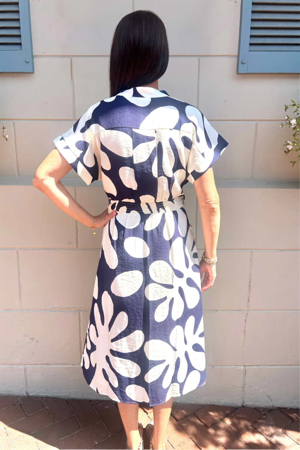 A woman viewed from behind wearing a short-sleeve belted midi dress with a bold navy and white floral pattern, standing against a light beige brick wall.