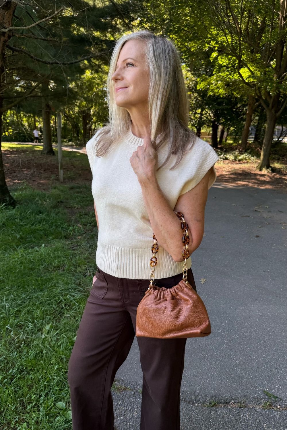 A woman standing outdoors on a park path wearing a cream ribbed sleeveless knit top, dark brown pants, and carrying a brown leather bucket bag with a gold chain strap.