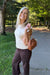 A smiling blonde woman stands outdoors on a paved path in a park, wearing a cream ribbed knit sweater and dark brown pants, while holding a brown leather shoulder bag with a gold chain strap.