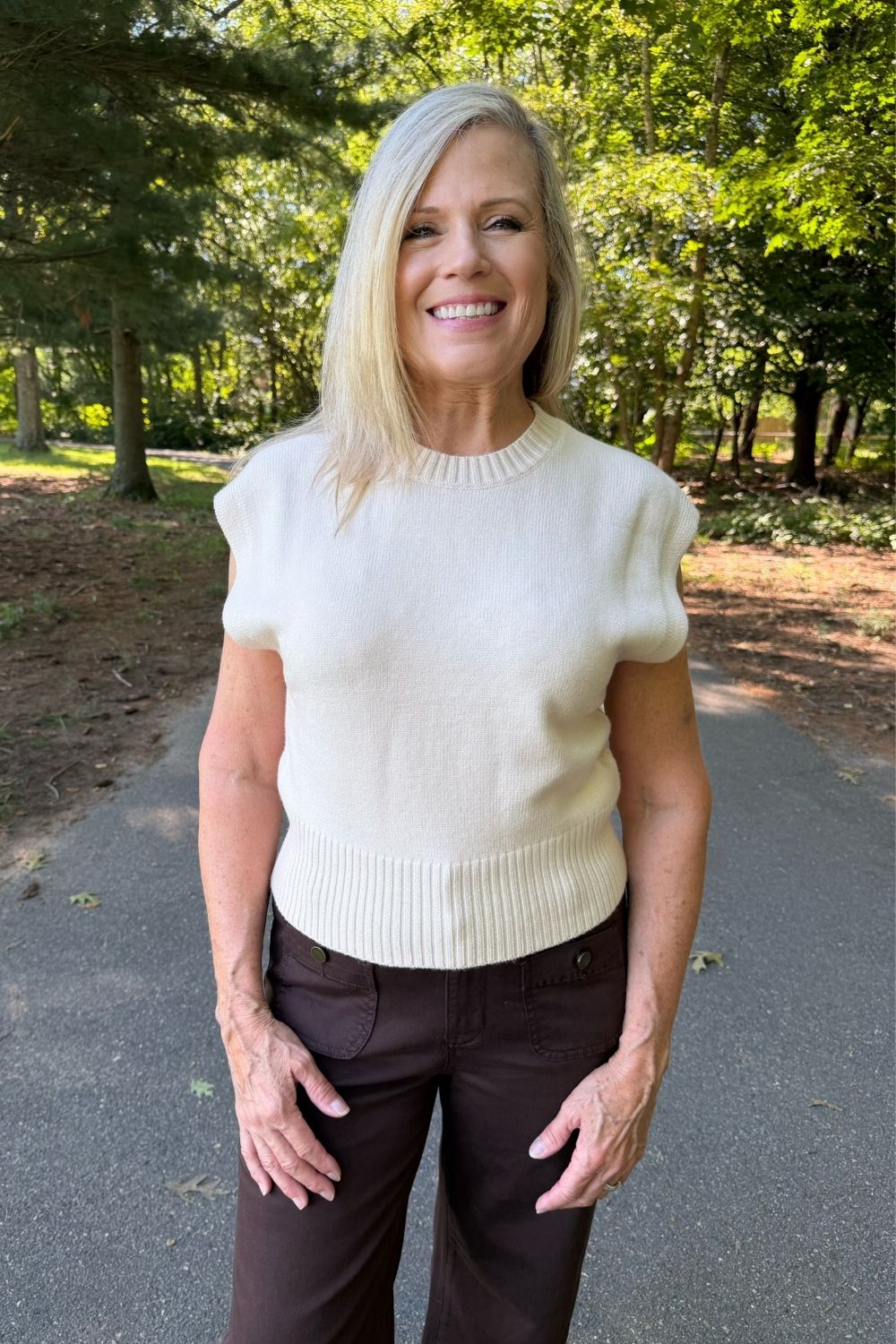 A smiling blonde woman wearing a cream-colored short-sleeve knit sweater and dark brown pants, standing outdoors on a paved path surrounded by green trees.