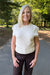 A smiling blonde woman wearing a cream-colored short-sleeve knit sweater and dark brown pants, standing outdoors on a paved path surrounded by green trees.