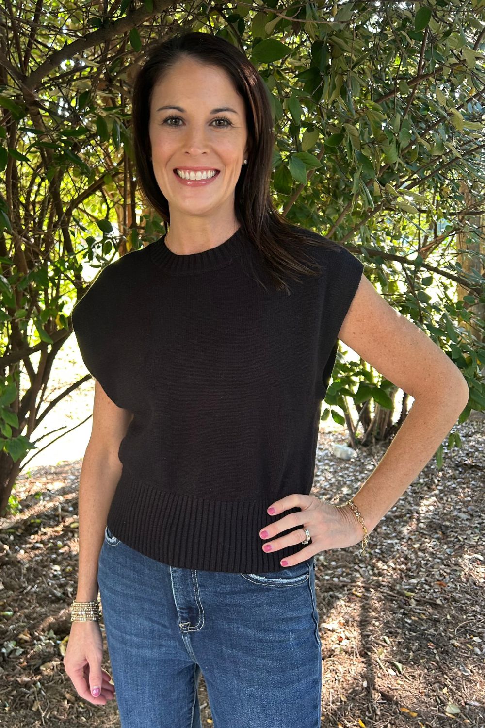 A woman wearing a black sleeveless ribbed knit top and blue jeans, standing outdoors in front of leafy trees.