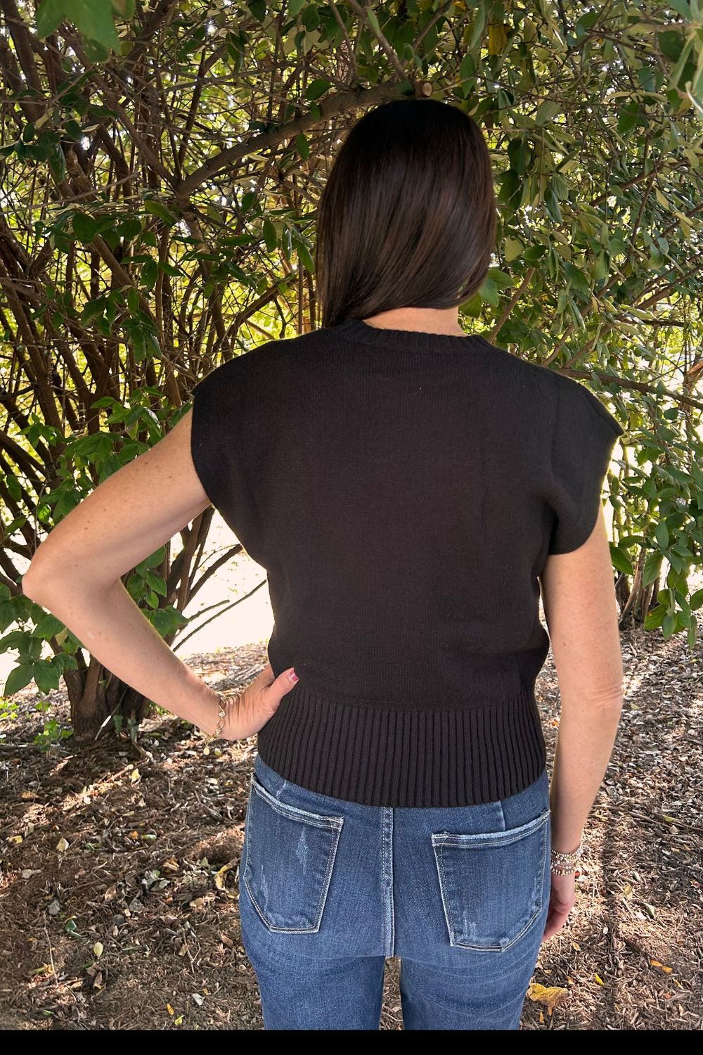 A woman viewed from behind wearing a black short-sleeve ribbed knit top and blue jeans, standing outdoors in front of leafy green bushes.