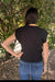 A woman viewed from behind wearing a black short-sleeve ribbed knit top and blue jeans, standing outdoors in front of leafy green bushes.