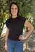 A woman wearing a black sleeveless ribbed knit top and blue jeans, standing outdoors in front of leafy trees.