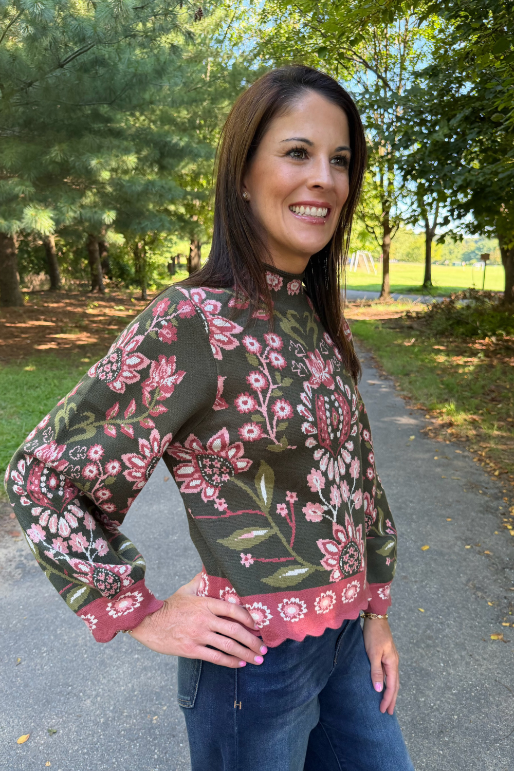 A woman wearing a dark olive green long-sleeve blouse with a pink and white floral pattern and scalloped pink cuffs, standing on a paved path in a park.