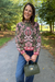A woman stands on a paved path in a park wearing a dark olive green mock-neck sweatshirt with a pink and red floral pattern, blue jeans, and holding an olive green handbag.