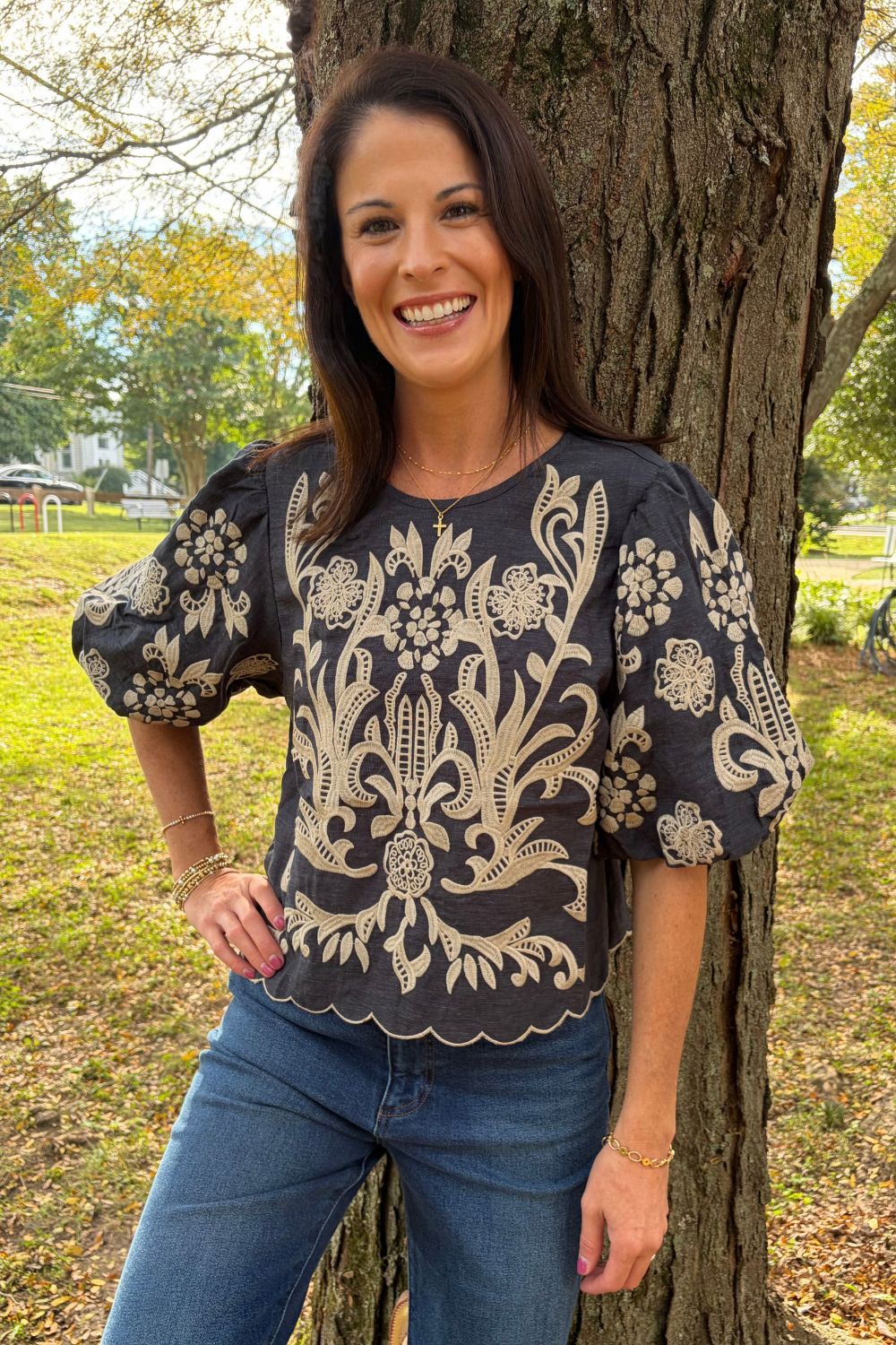 A woman smiling outdoors next to a tree, wearing a dark charcoal gray short-sleeve blouse with beige floral embroidery and blue jeans.