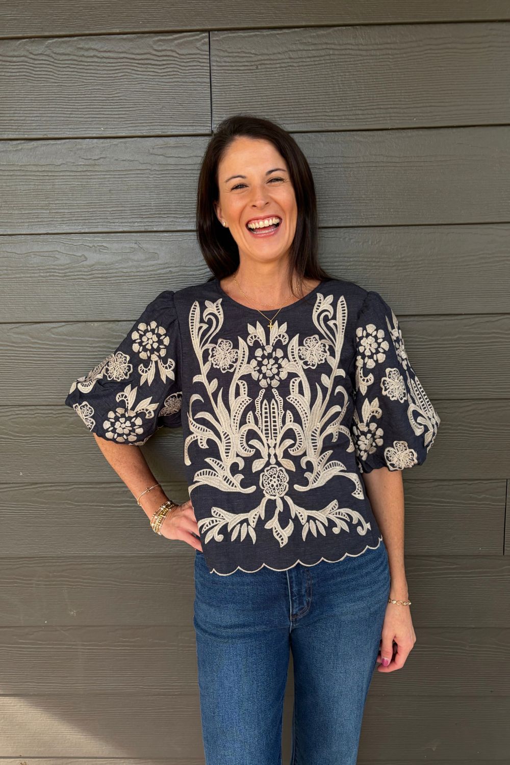 A woman smiling and wearing a navy blue blouse with intricate white floral embroidery and puff sleeves, paired with blue jeans, standing against a gray wood-paneled wall.