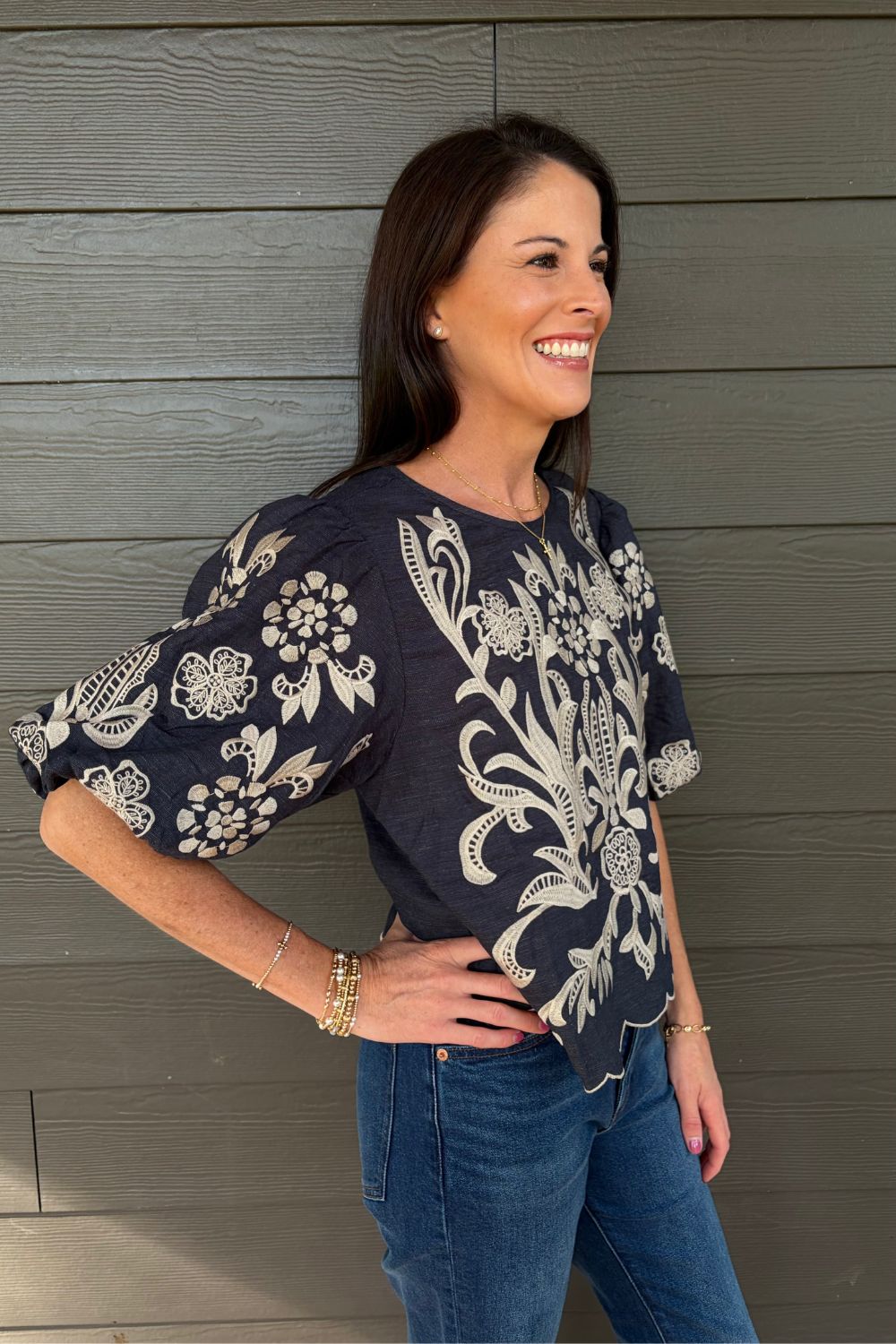 A woman wearing a navy blue short-sleeve blouse with intricate white floral embroidery, paired with blue jeans, standing against a gray wood-paneled wall.