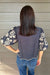 A woman shown from the back wearing a navy blue short-sleeve blouse with intricate white floral embroidery, paired with blue jeans, standing against a gray wood-paneled wall.