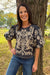 A woman smiling outdoors next to a tree, wearing a dark charcoal gray short-sleeve blouse with beige floral embroidery and blue jeans.