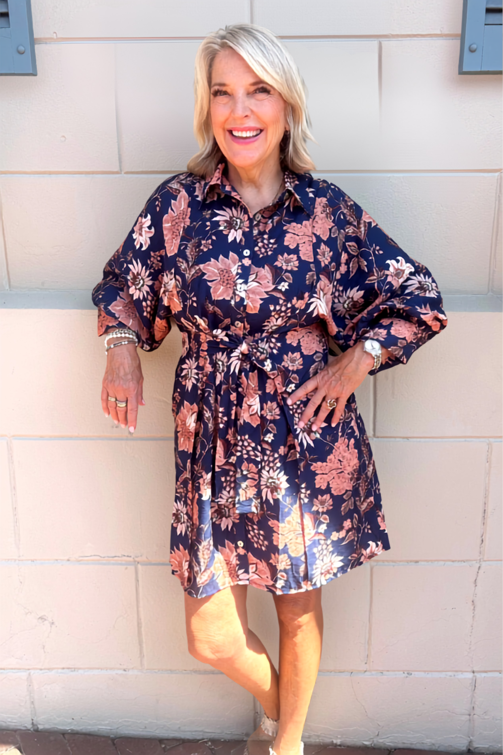 A smiling blonde woman stands against a light beige exterior wall wearing a navy floral shirt dress with pink, peach, and white flowers. The dress has a button-up collar, long sleeves, and a self-tie waist belt.