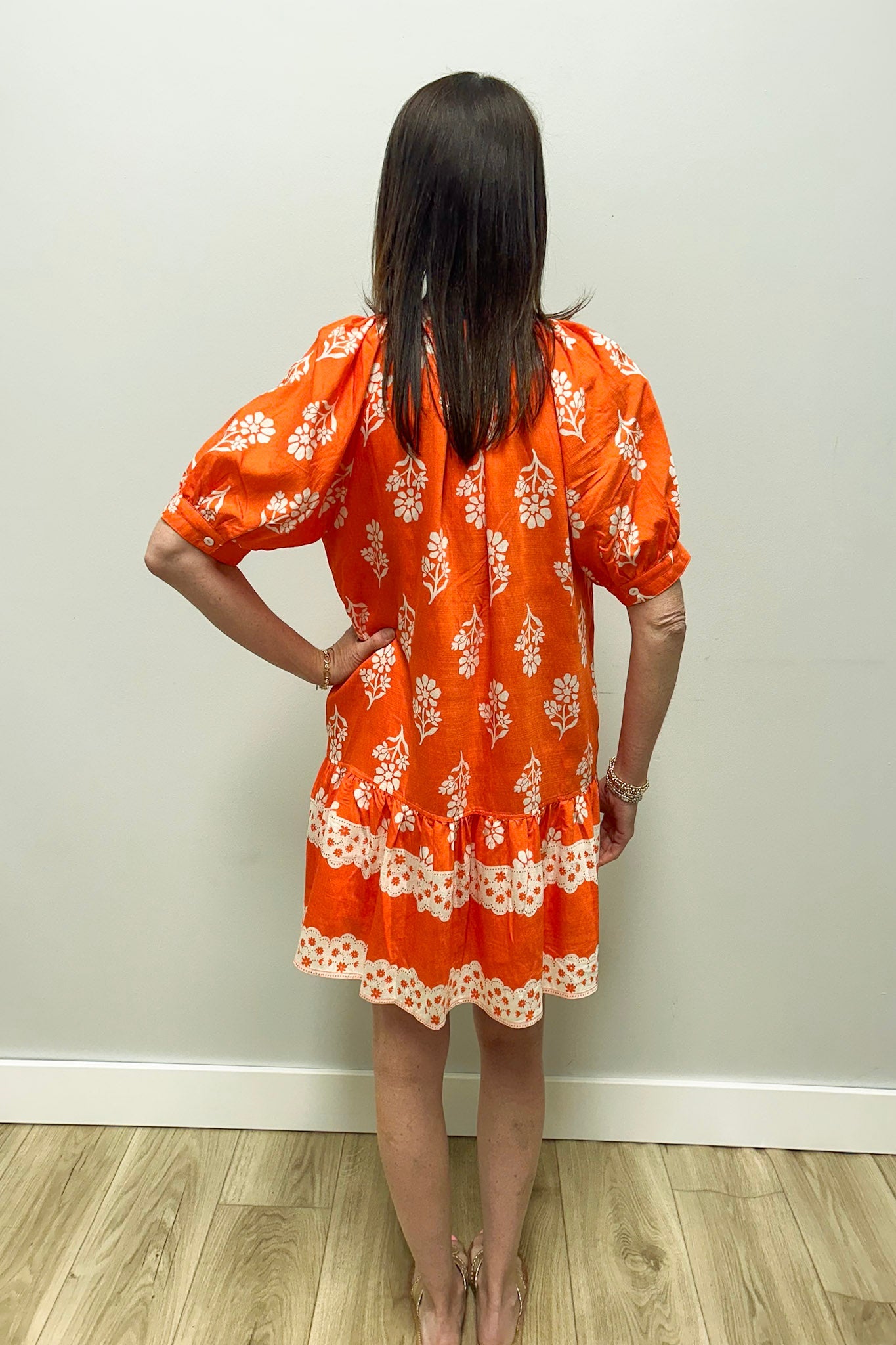 Woman wearing an orange dress with white floral pattern against a plain wall.