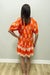 Woman wearing an orange dress with white floral pattern against a plain wall.