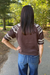 A woman viewed from behind wearing a brown short-sleeve knit sweater with cream geometric patterns on the shoulders and sleeves, paired with blue jeans, standing on a paved path in a wooded outdoor setting.