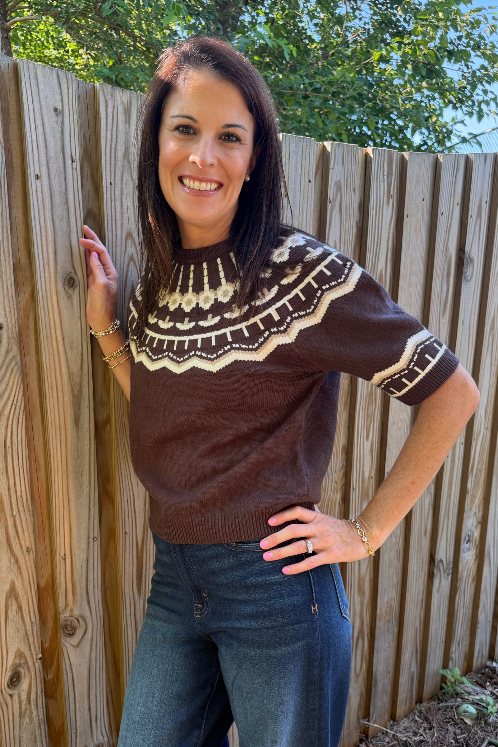 A woman wearing a brown short-sleeve knit sweater with a cream geometric pattern around the yoke and sleeve cuffs, standing outdoors against a wooden fence.