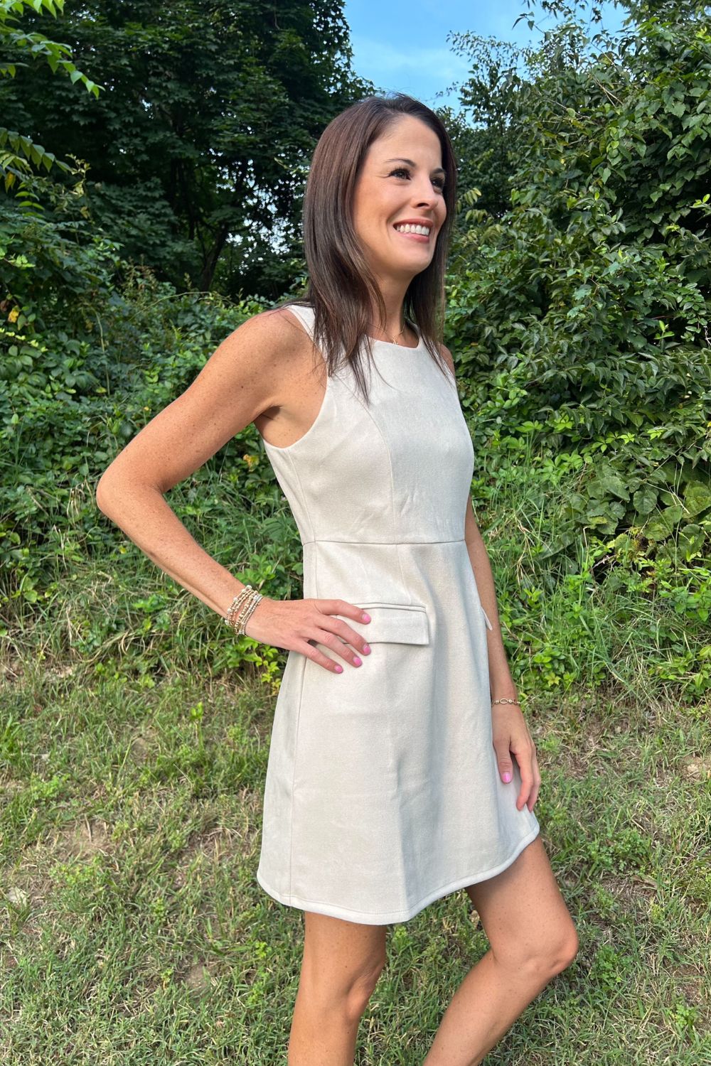 A woman wearing a sleeveless light beige linen dress with a front patch pocket, standing outdoors in a grassy area with green foliage and blue sky in the background.