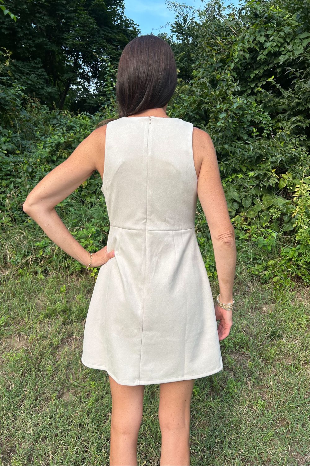 A woman viewed from behind wearing a sleeveless, light beige or off-white mini dress with a structured, slightly flared silhouette, standing outdoors on grass with green foliage in the background.