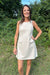 A woman wearing a sleeveless light beige linen dress with a front patch pocket, standing outdoors in a grassy area with green foliage and blue sky in the background.