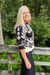 A woman with blonde hair stands on a wooden boardwalk outdoors, wearing a black and white patterned short-sleeve top and dark blue jeans, with green trees in the background.