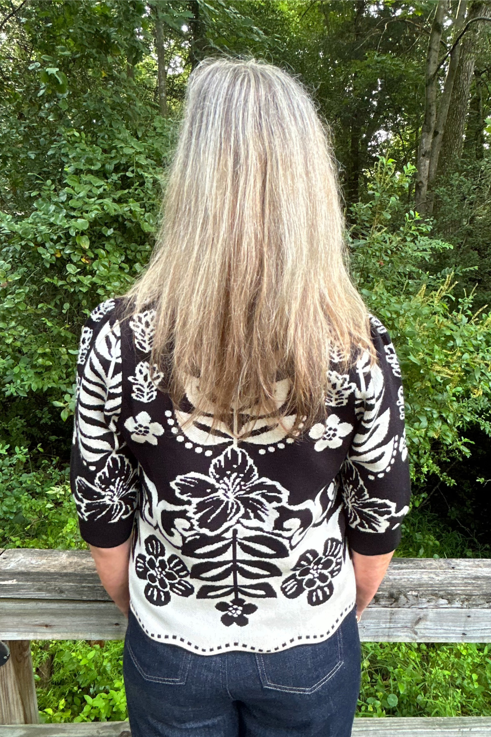 A woman viewed from behind wearing a black and white floral patterned knit top, standing outdoors in front of a wooden fence and green trees.