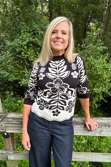 A smiling blonde woman stands outdoors in front of a wooden fence and green trees, wearing a black and cream knit sweater with a floral and leaf pattern and dark blue jeans.