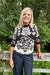 A smiling blonde woman stands outdoors in front of a wooden fence and green trees, wearing a black and cream knit sweater with a floral and leaf pattern and dark blue jeans.