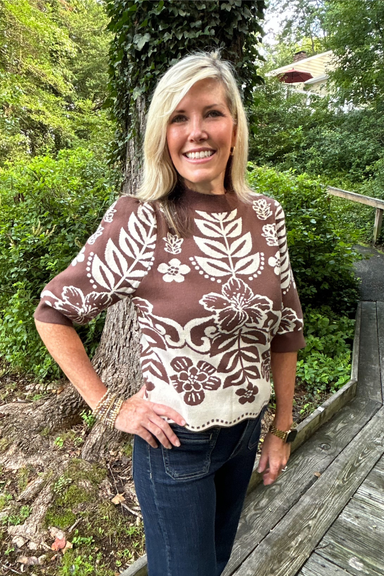 A smiling blonde woman wearing a brown and cream patterned short-sleeve top and dark blue jeans, standing outdoors on a wooden boardwalk with green foliage and a tree trunk in the background.