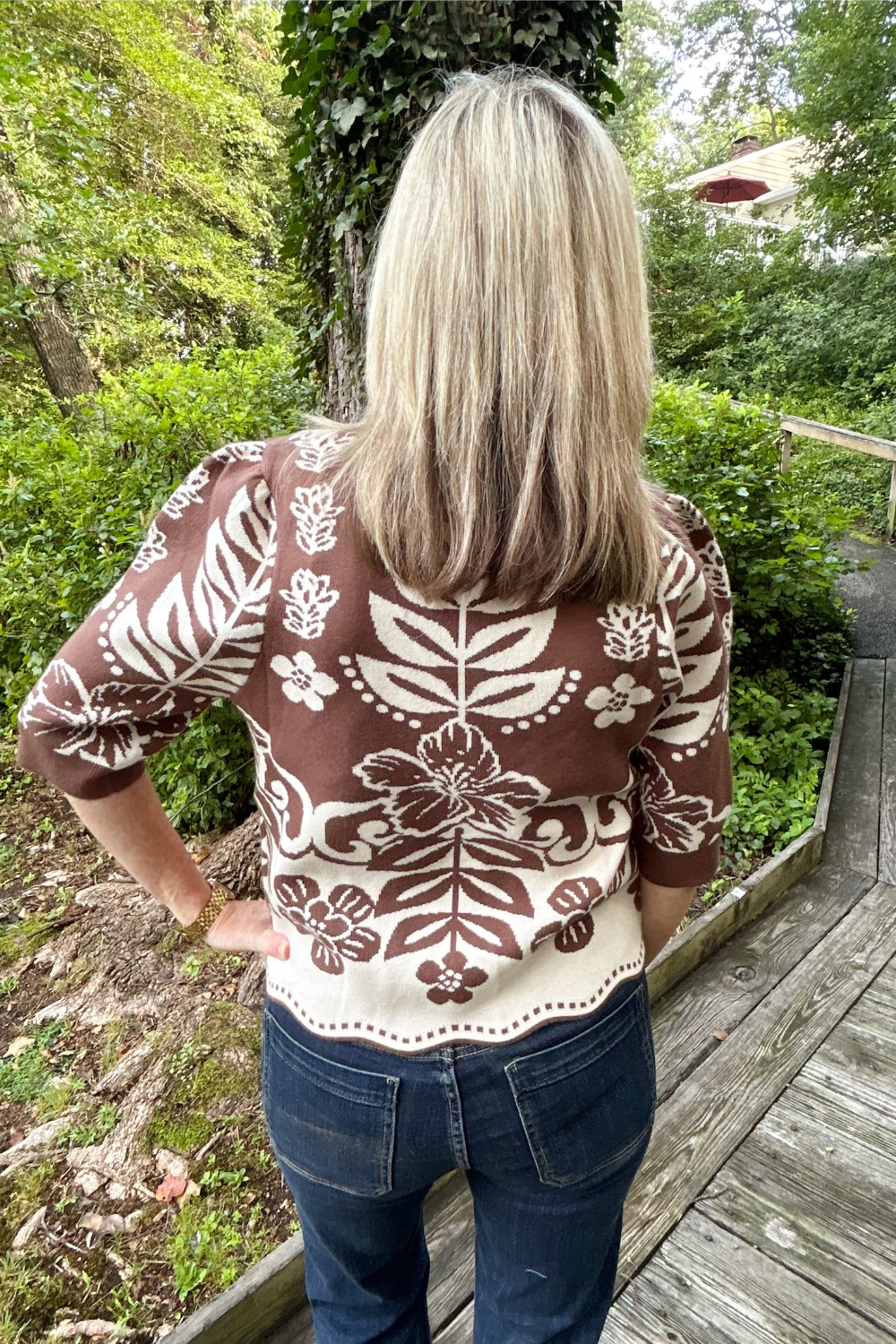 A woman viewed from behind wearing a brown and cream floral patterned knit top and blue jeans, standing on a wooden boardwalk surrounded by green trees and foliage.
