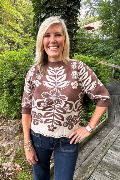 A smiling blonde woman wearing a brown and cream floral patterned knit sweater and dark blue jeans, standing outdoors on a wooden boardwalk surrounded by green foliage.