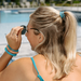 Woman with blonde hair in a ponytail wearing sunglasses and turquoise accessories by a pool.