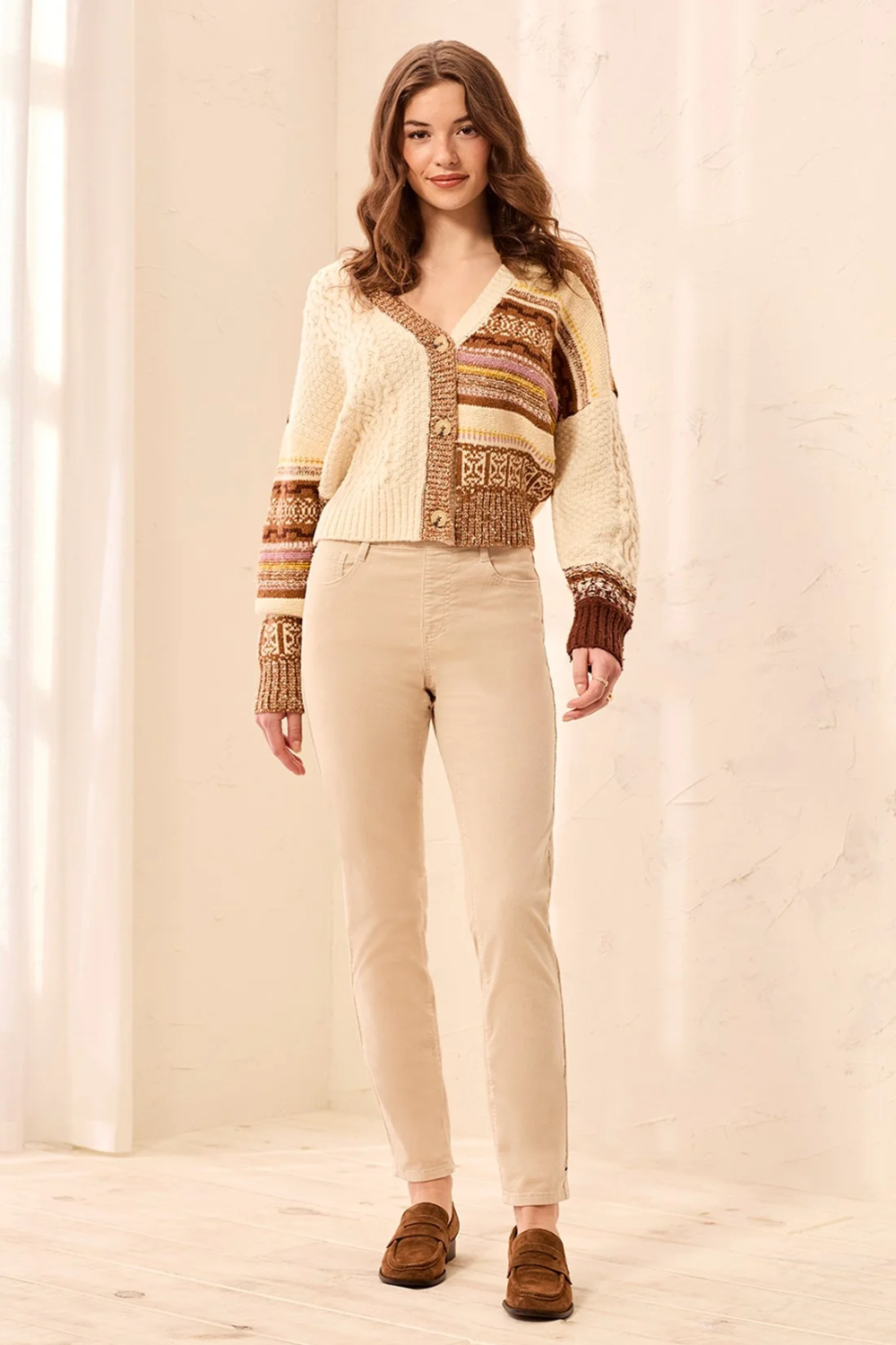 A woman modeling a cream and brown knit cardigan with a Fair Isle patterned chest panel and striped cuffs, paired with beige pants and brown suede loafers, in a bright indoor setting.