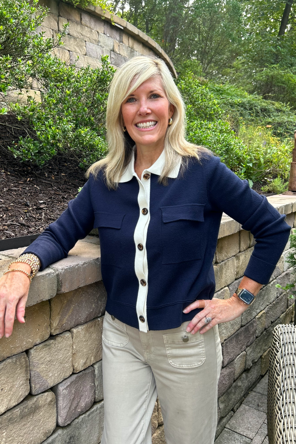 A smiling blonde woman stands outdoors against a stone wall and greenery, wearing a navy cardigan with a white collar and front buttons, paired with khaki pants.