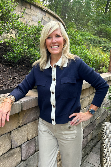 A smiling blonde woman stands outdoors against a stone wall and greenery, wearing a navy cardigan with a white collar and front buttons, paired with khaki pants.
