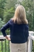 A woman viewed from behind wearing a navy blue knit sweater and beige pants, standing on a white railing with a lush green forest background.