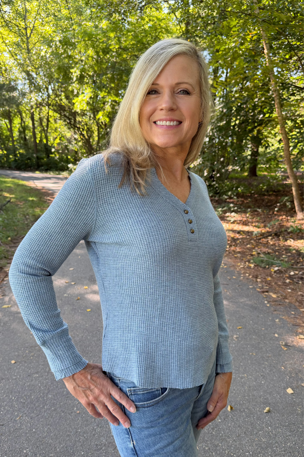 A smiling blonde woman wearing a light blue-gray ribbed long-sleeve henley shirt with three buttons, paired with blue jeans, standing outdoors on a paved path surrounded by green trees.