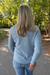 A woman viewed from behind wearing a light blue textured knit sweater and blue jeans, standing on a paved path in a wooded outdoor setting.