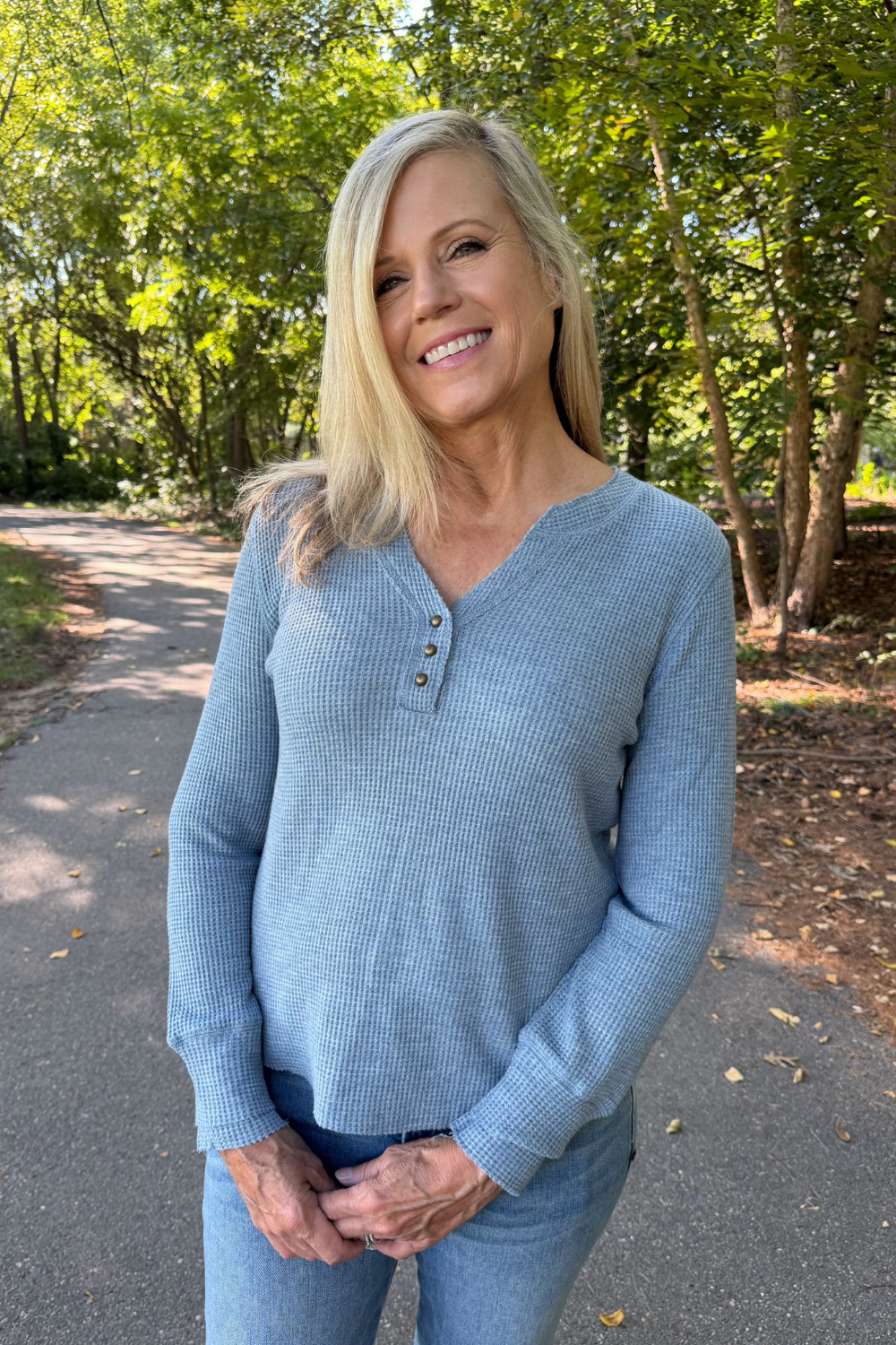 A smiling blonde woman wearing a light blue ribbed long-sleeve henley sweater and blue jeans, standing on an outdoor paved path surrounded by green trees.