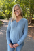 A smiling blonde woman wearing a light blue ribbed long-sleeve henley sweater and blue jeans, standing on an outdoor paved path surrounded by green trees.
