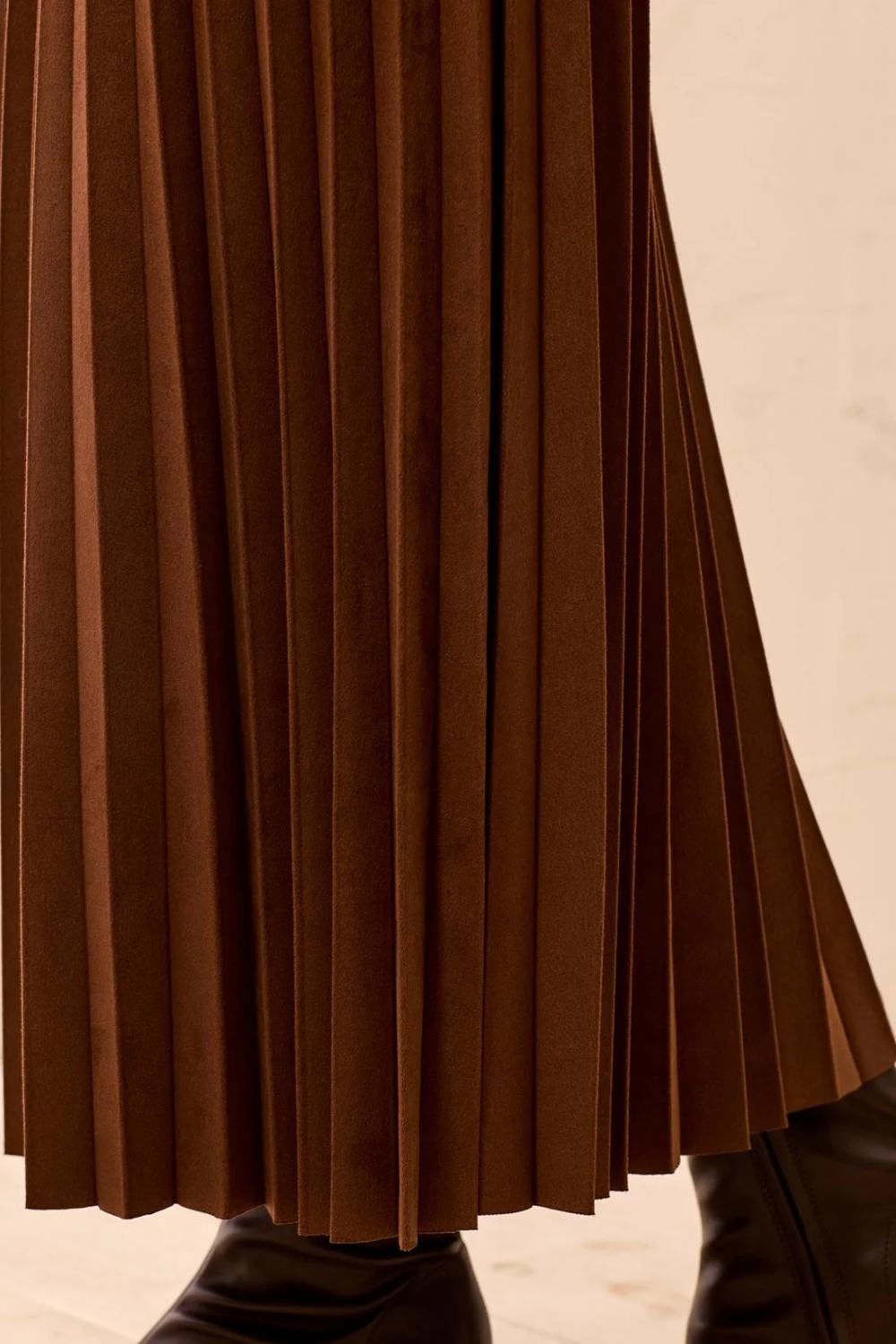 Close-up of a brown pleated skirt with a soft suede-like texture, paired with black boots against a light beige background.