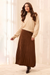 A woman modeling a cream knit mock-neck sweater and a brown pleated midi skirt, standing in a bright, neutral indoor setting with sheer curtains.